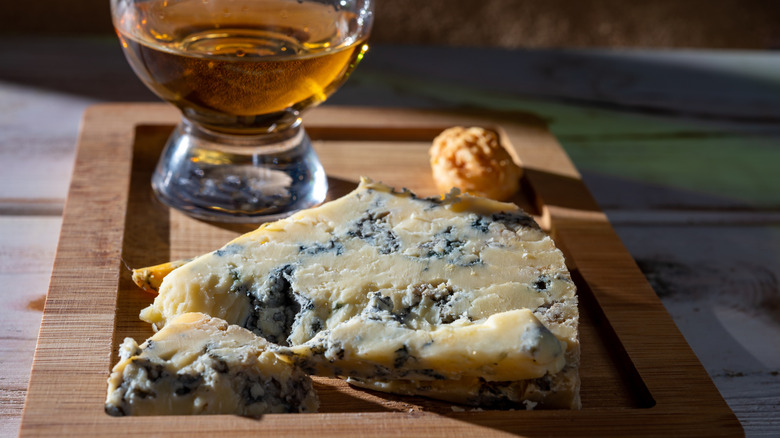 A piece of blue cheese on a wooden board with a glass of bourbon