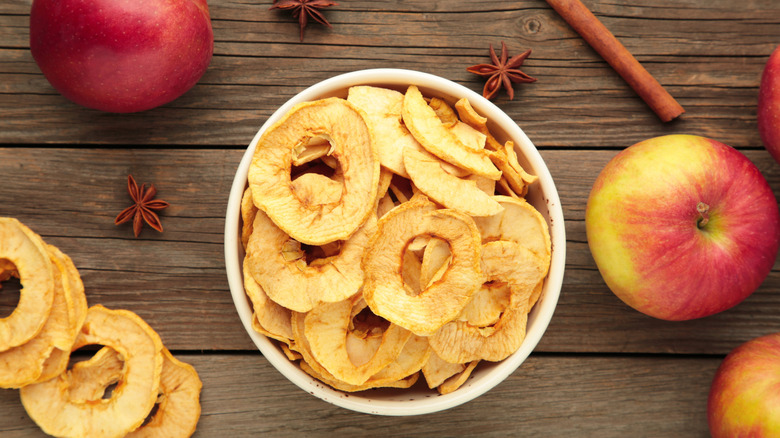 A bowl of dried apple chips next to whole fresh apples and cinnamon sticks