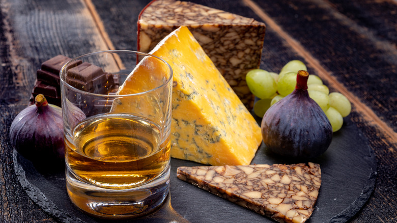 A glass of whisky on a plate with cheese, grapes, figs, and dark chocolate