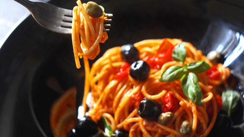 A fork full of pasta puttanesca, with the bowl of pasta in the background
