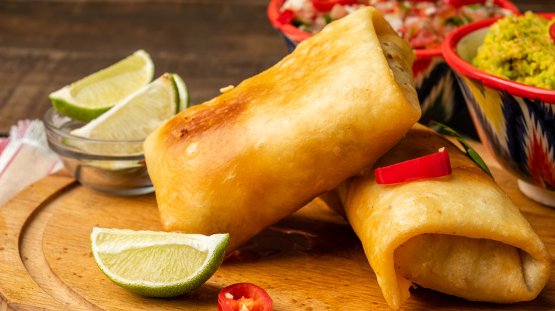 Two chimichangas sit on a wooden cutting board with guacamole and limes on the side