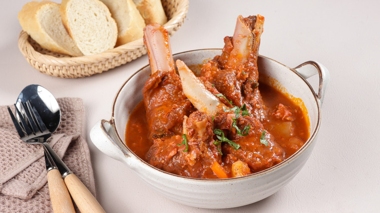 A bowl of tulang merah on a table with cutlery and a basket of bread