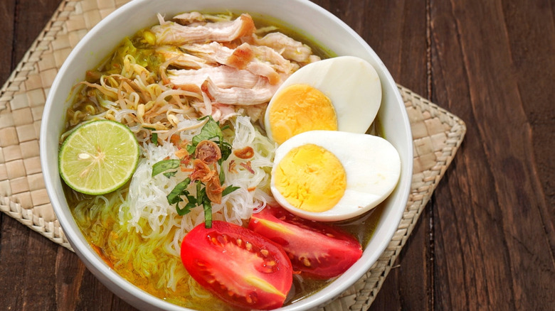 A bowl of Indonesian soto ayam with chicken, hard boiled eggs, tomatoes, noodles, and lime