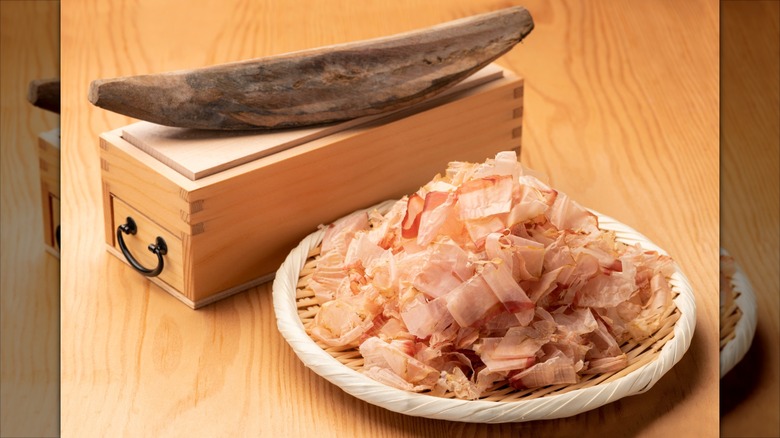 A piece of katsuobushi skipjack tuna sitting on top of a kezuriki mandoline box next to a plate of bonito flakes