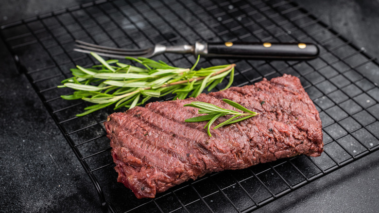 A grilled flank steak resting on a rack with rosemary garnish