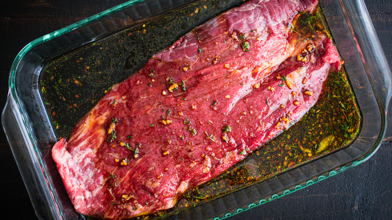 A raw flank steak with marinade in a glass baking dish