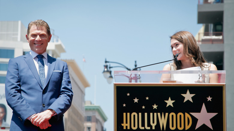 Sophie Flay talks into a microphone about her dad, Bobby Flay, who stands nearby