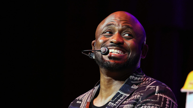 Wayne Brady smiling on stage with a clip on microphone