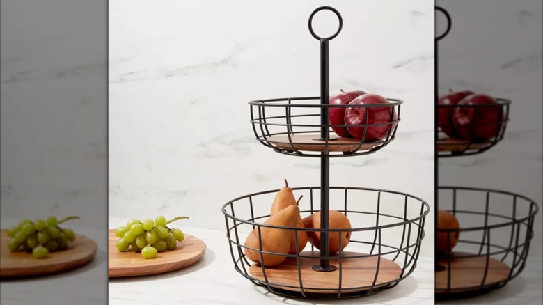 Wood and metal fruit basket with various fruits inside next to wood cutting board.