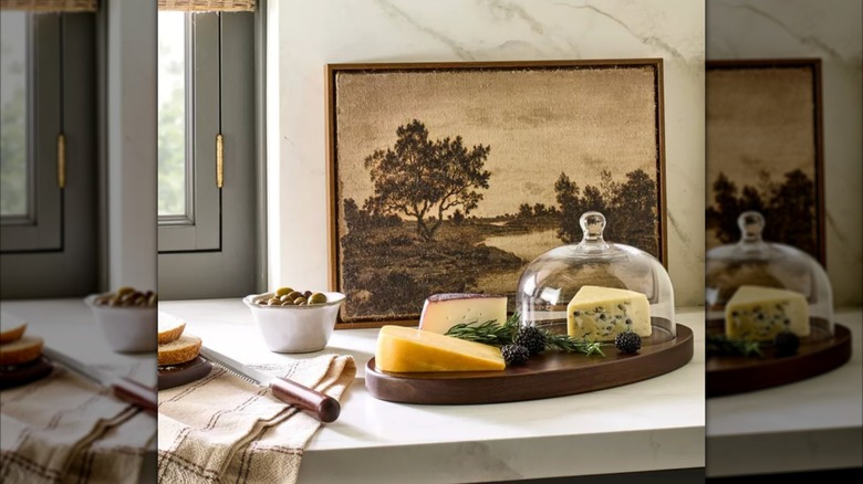 Target wood cheese board with cloche on counter in front of nature painting.