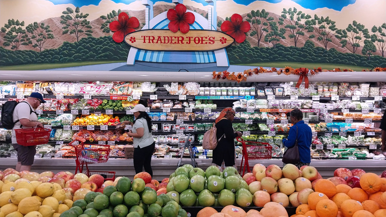 Interior of Trader Joe's showing four people shopping by the refrigerated shelves in the produce section with a row of produce in front of them