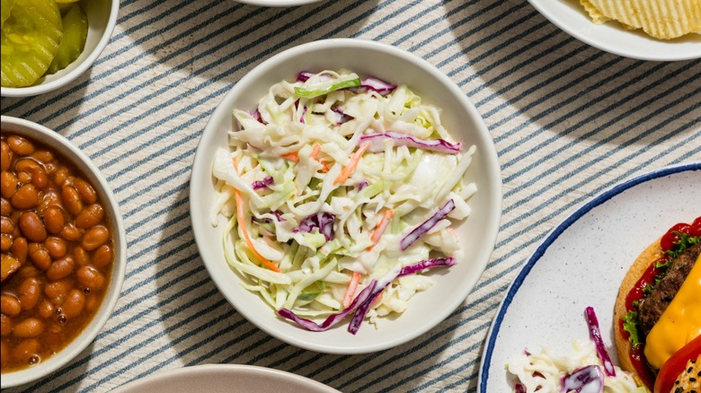 A bowl of creamy coleslaw surrounded by other barbecue food and side dishes on a striped tablecloth