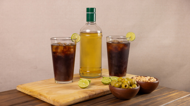 Piscola cocktails on a cutting board with a bottle of Pisco and small bowls of olives and nuts