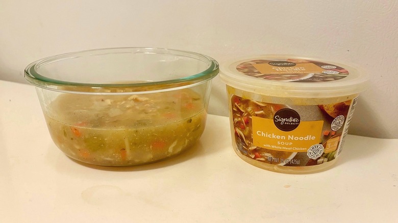 A tub of Signature Select Chicken Noodle Soup next to a prepared bowl of soup