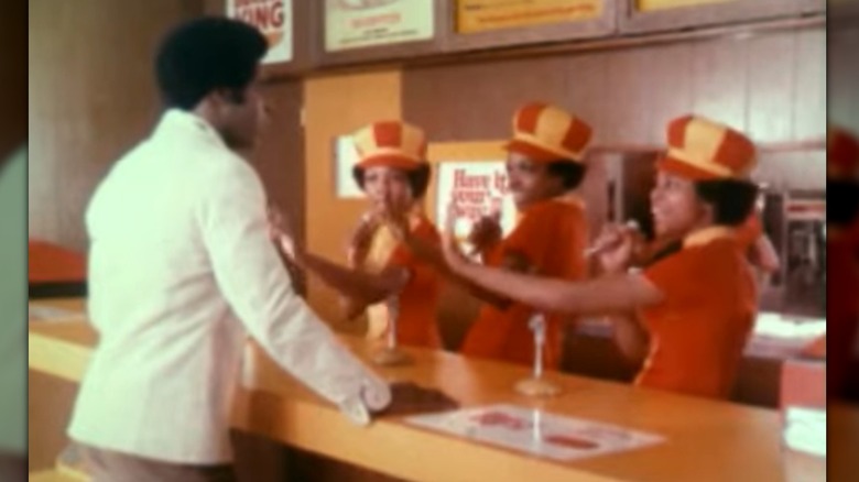 Still showing a man in a white jacket at a counter while three women in orange and yellow uniforms sing and dance in unison