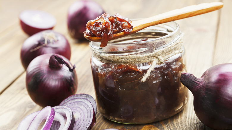 Red onion jam in a glass jar with a wooden spoon and fresh onions surrounding it