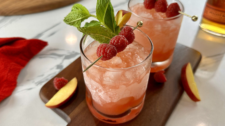 two peach and raspberry bourbon smashes on wooden cutting board