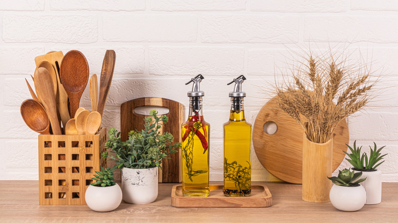 Two glass bottles of cooking oil surrounded by decorative plants, a cutting board, and a crock full of wooden cooking tools