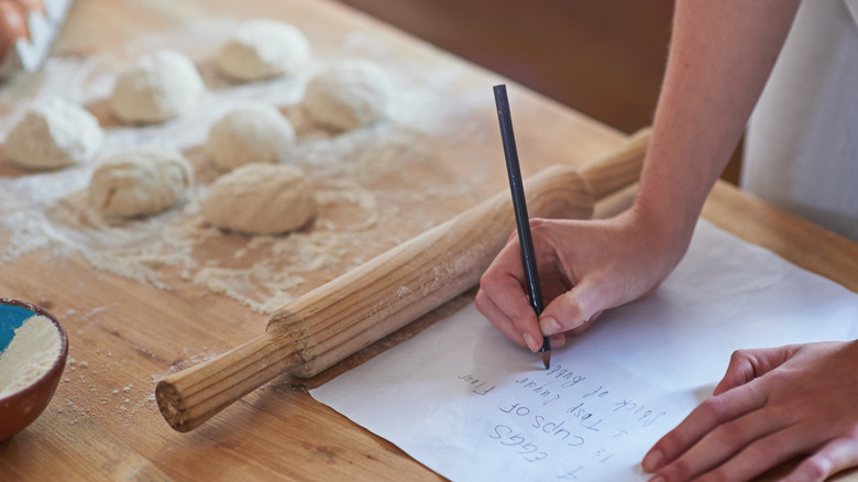 Person writing recipe instructions on paper beside rolling pin and dough