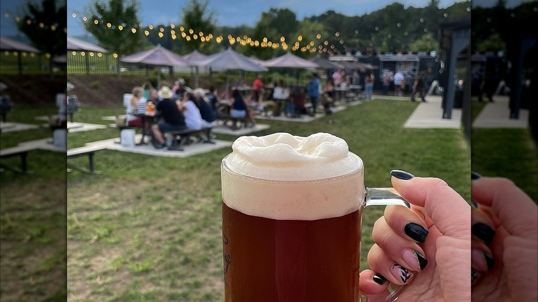 A person holding a frothy beer with a beer garden in the background at dusk