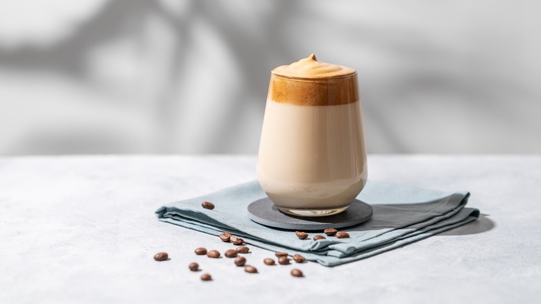 A clear glass with dalgona coffee in it, on a napkin with coffee beans spread around it