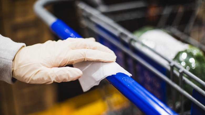 A nitrile gloved hand uses a sanitary wipe on the handle of a shopping cart