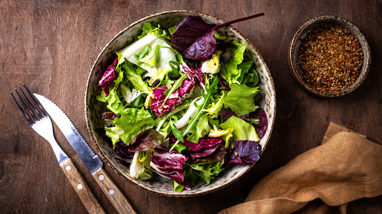 Fresh mixed greens salad in ceramic bowl next to cutlery on wood table.