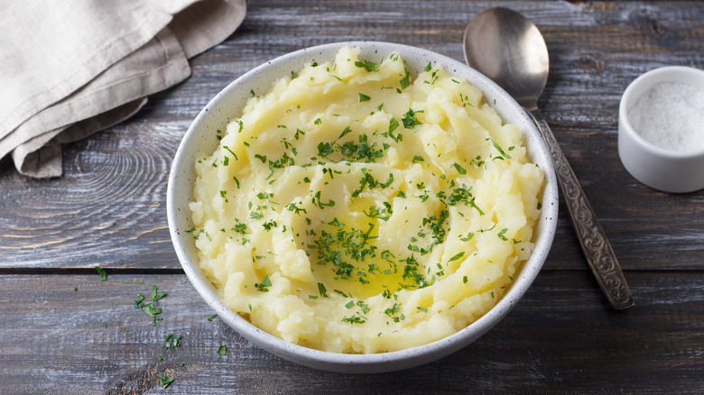 Herby mashed potatoes in white bowl next to spoon on dark gray wood table.