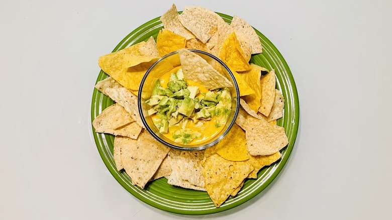 A green plate has tortilla chips and a bowl of queso with avocado.