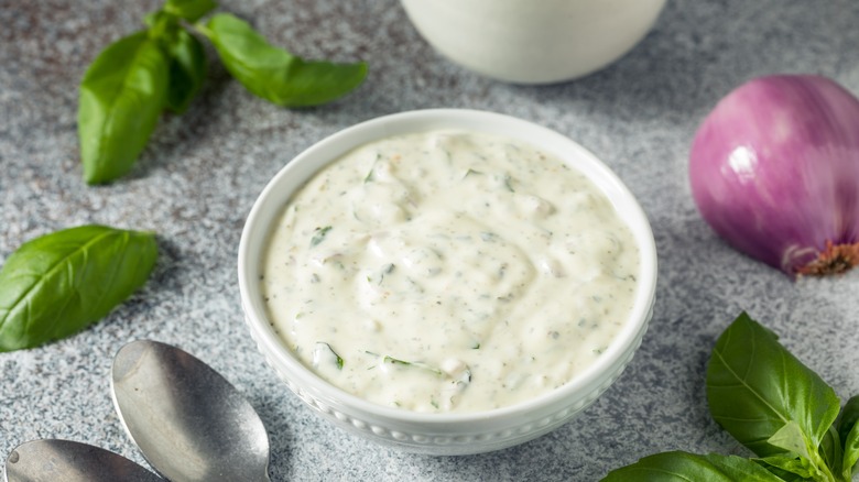 Green goddess dressing in white bowl with basil and a red onion in the background