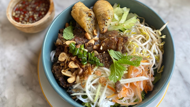 A bowl of bún thịt nướng topped with fresh herbs, peanuts, and spring rolls