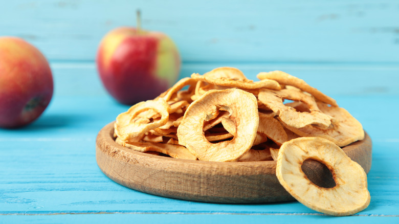 Pile of apple chips on a wooden plate with two apples in the background