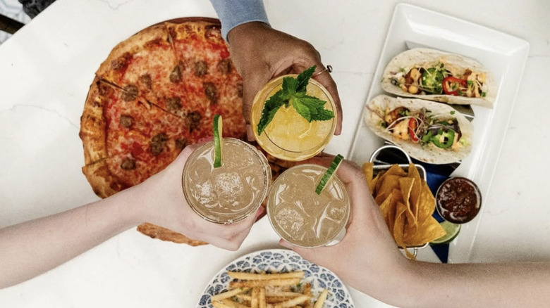 An overhead shot of three people holding cocktails over a table spread with food