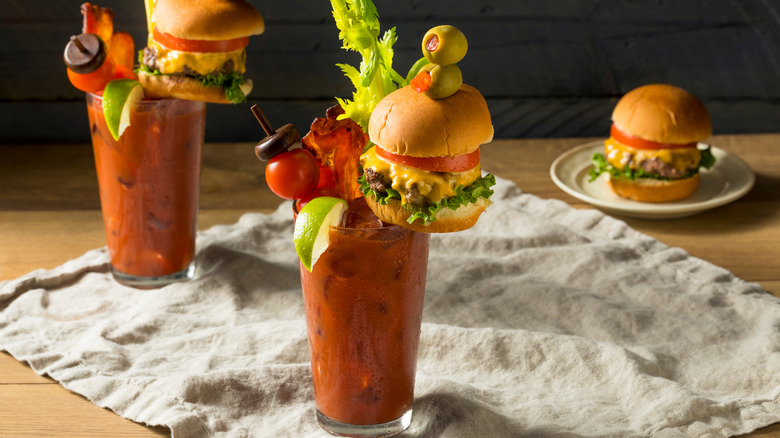 A loaded Bloody Mary on a table garnished with a cheeseburger, olives, and lime, with another drink and a cheeseburger in the background