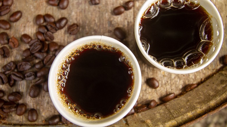 Two cups of Kona coffee on a wooden surface surrounded by coffee beans