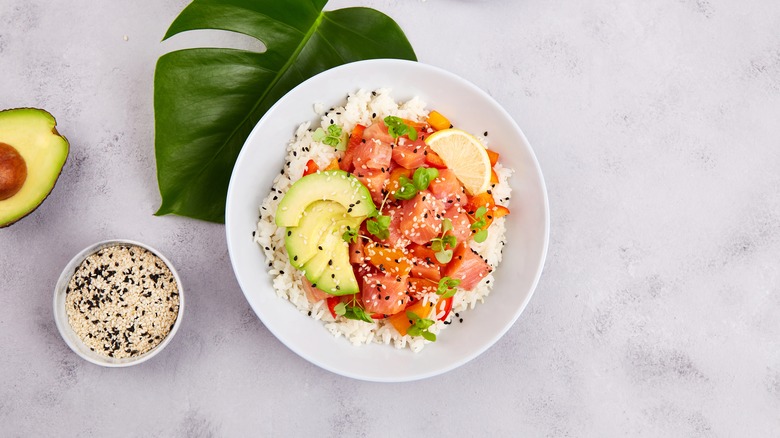 A bowl of salmon poke topped with avocado next to a bowl of chia seeds on a white countertop