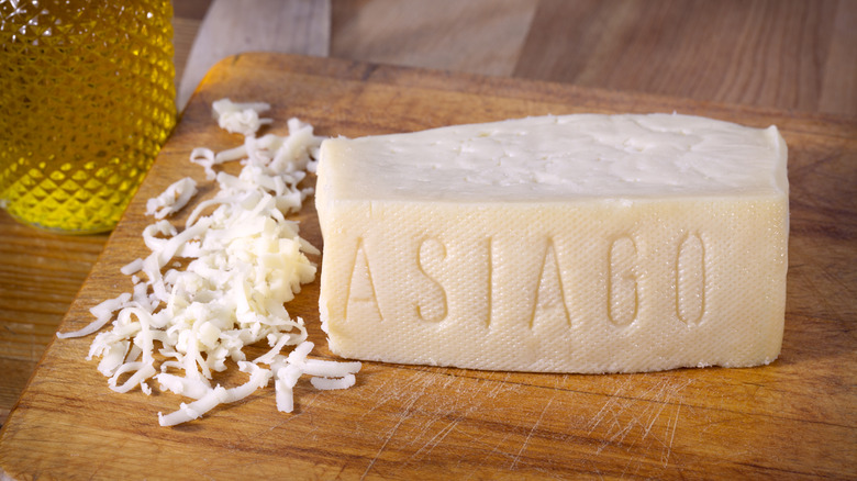 A wedge of Asiago cheese on a wooden board next to a pile of shredded Asiago.