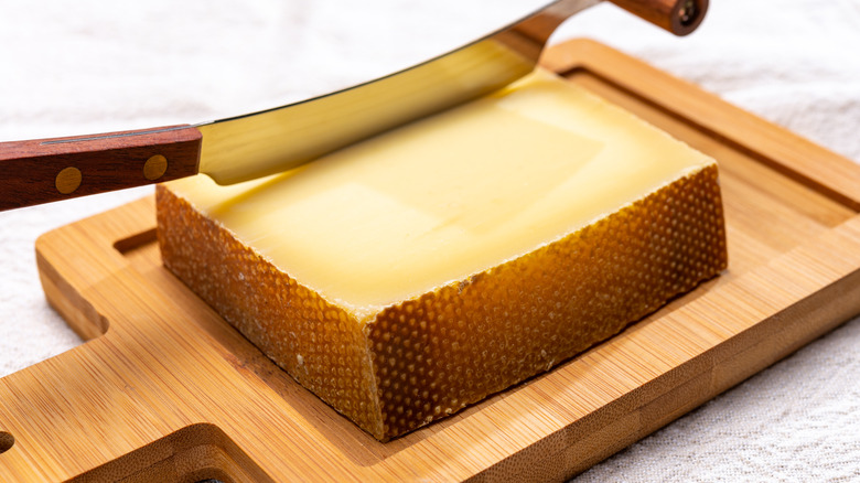 A thick wedge of Gruyère cheese sliced by a knife on a wooden cutting board.