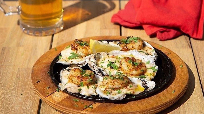 A platter of oysters topped with a seared shrimp on a wooden table with a frothy beer in the background