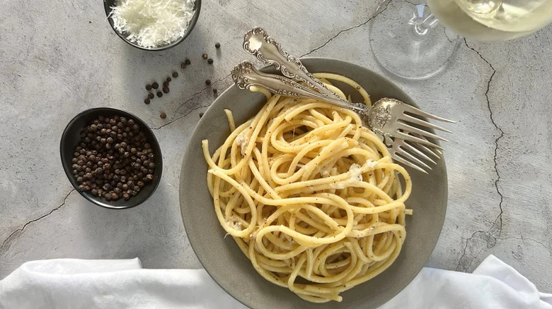 Grey bowl with cacio e pepe pasta in it, ramekin of black peppercorns, ramekin of fresh grated parmesan, and glass of white wine