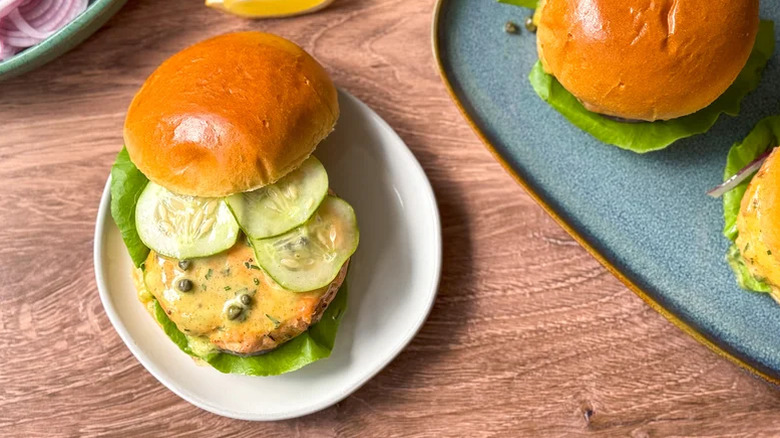 White plate with a salmon burger on a bun garnished with sliced cucumber, lettuce, and curry remoulade sauce