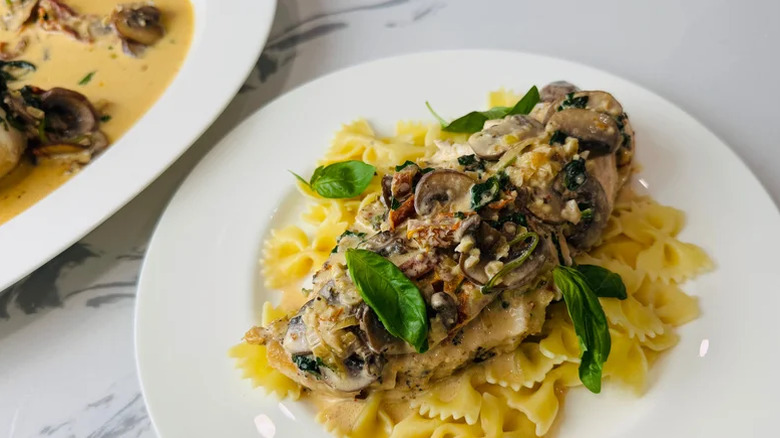White plate of chicken breast with sun-dried tomato, spinach, and mushroom sauce over a bed of farfalle