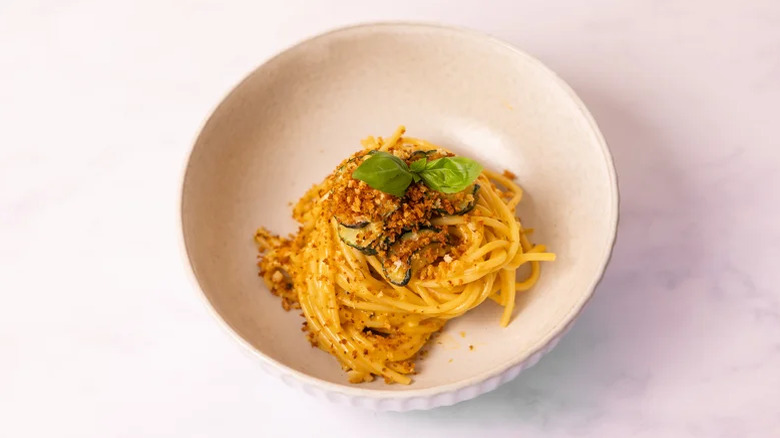 White bowl of vegetarian spaghetti 'carbonara' garnished with basil, toasted panko bread crumbs, and slices of zucchini
