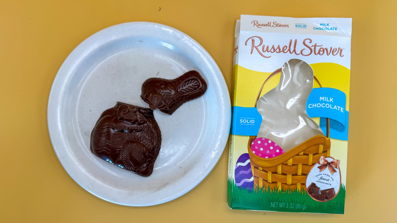 Russell Stover Easter bunny chocolate broken in two at the neck on a white plate next to packaging on a yellow background