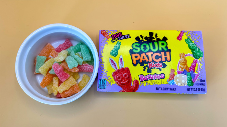 Pastel-colored Sour Patch Kids Bunnies candy in a white ramekin next to a box of the candy on a yellow background