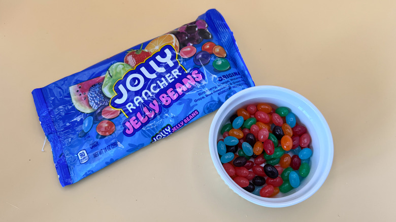 Multi-colored Jolly Rancher jellybeans in a white ramekin next to packaging on a yellow background
