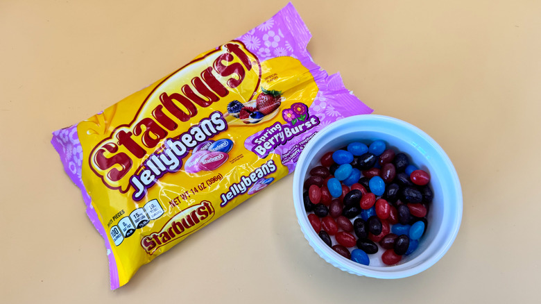 Red, dark purple, and blue Starburst jellybeans in ramekin next to a package of the candy on a yellow background