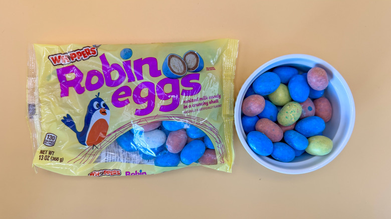 Whoppers Robin Eggs in a package next to a white ramekin full of red, blue, and yellow speckled eggs on yellow background