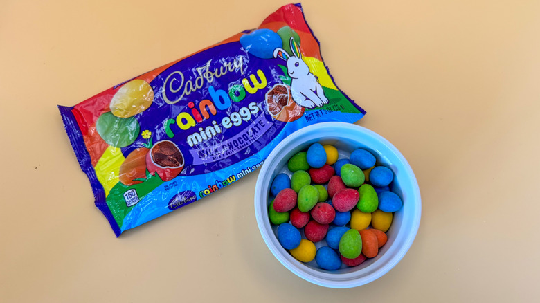 Multicolored mini Cadbury eggs in a white ramekin next to packaging on a yellow background