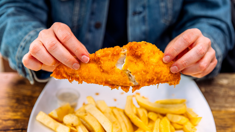 Someone breaking apart a fried fish fillet above a plate of fries at an outdoor table